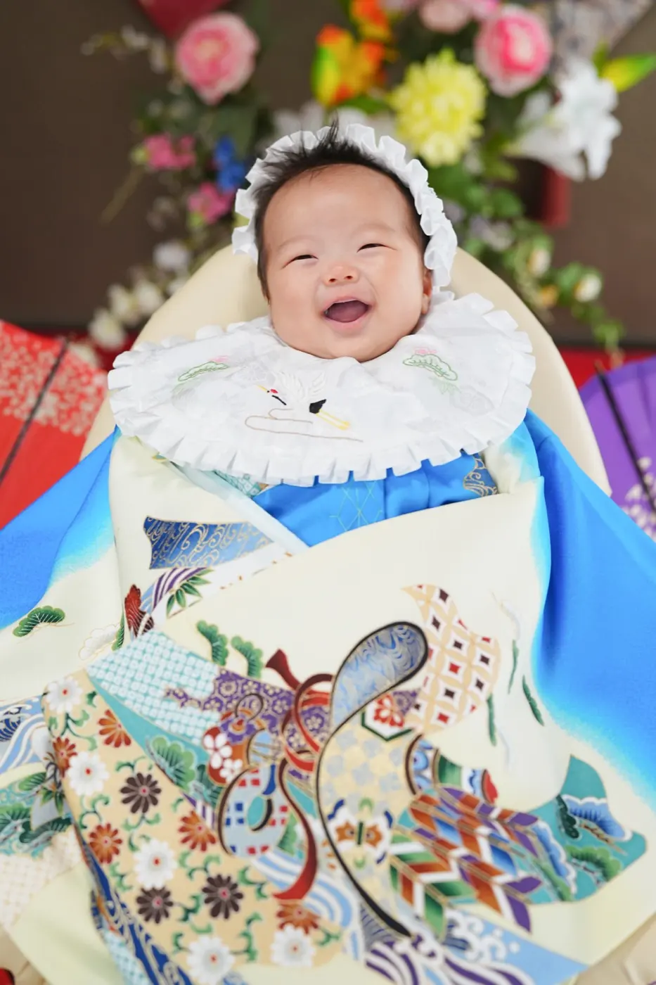 青い祝い着をかけて、白いフリルの帽子とよだれかけで大きく笑うお宮参りの赤ちゃん