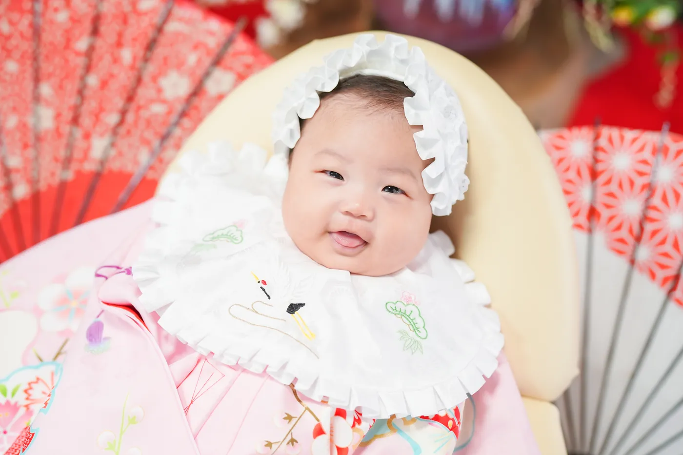 赤い和傘の前で、白いボンネットをかぶった祝い着の赤ちゃん