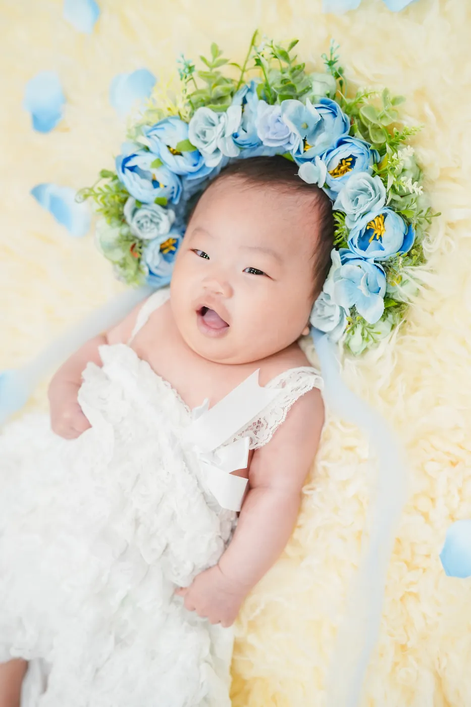 青いお花の冠をつけて、白いふわふわの上で笑う赤ちゃん