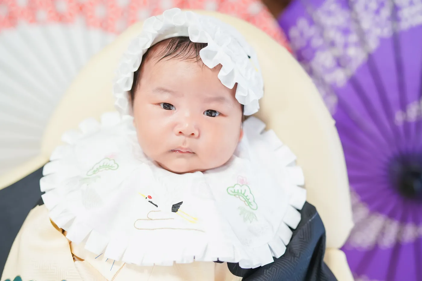 紫の和傘の前で、白いボンネットをかぶった赤ちゃん