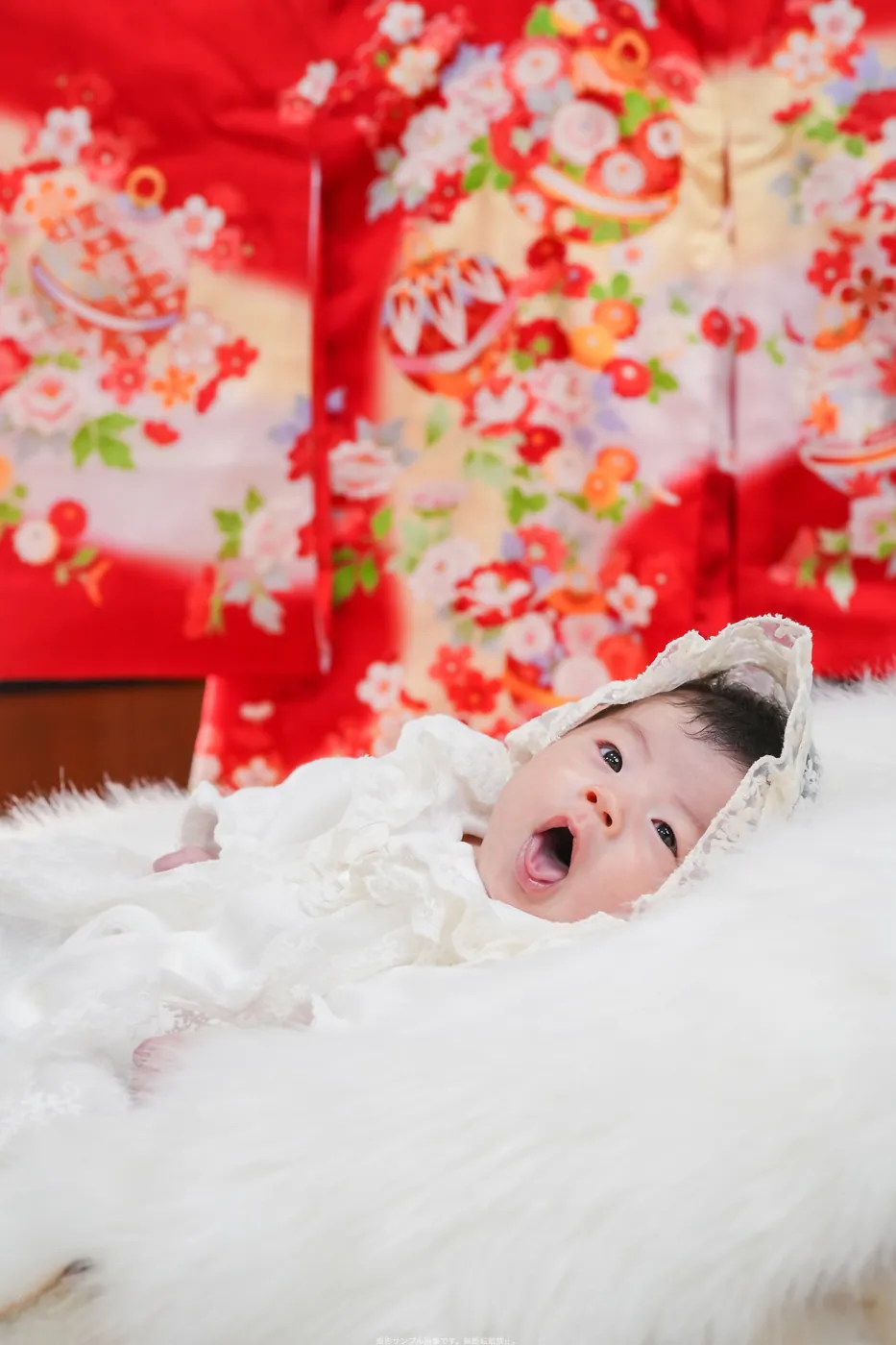 赤い祝い着を背に、ふわふわの上であくびする赤ちゃん