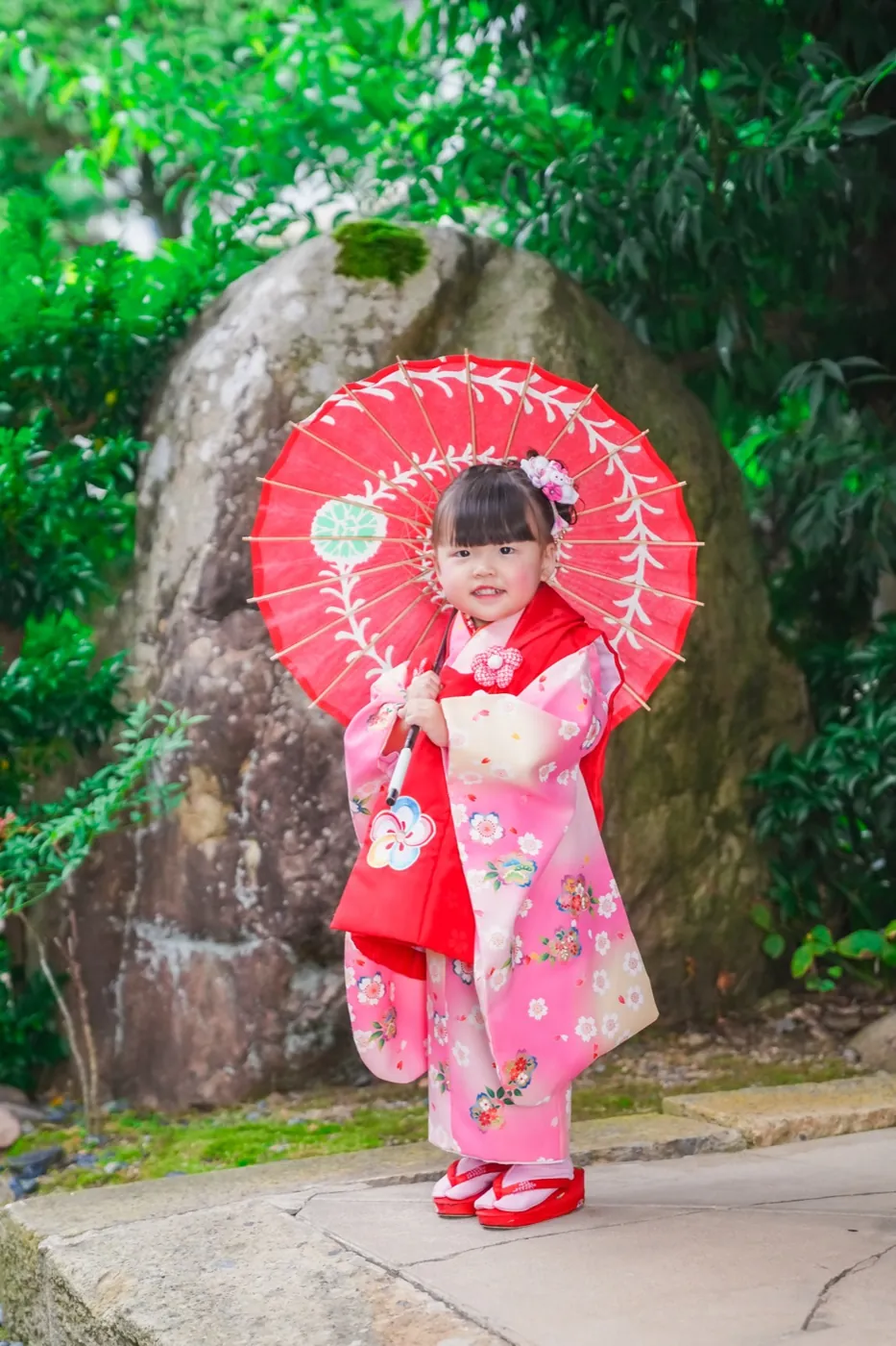 庭の大きな岩の前で赤い和傘を持つ3歳の女の子