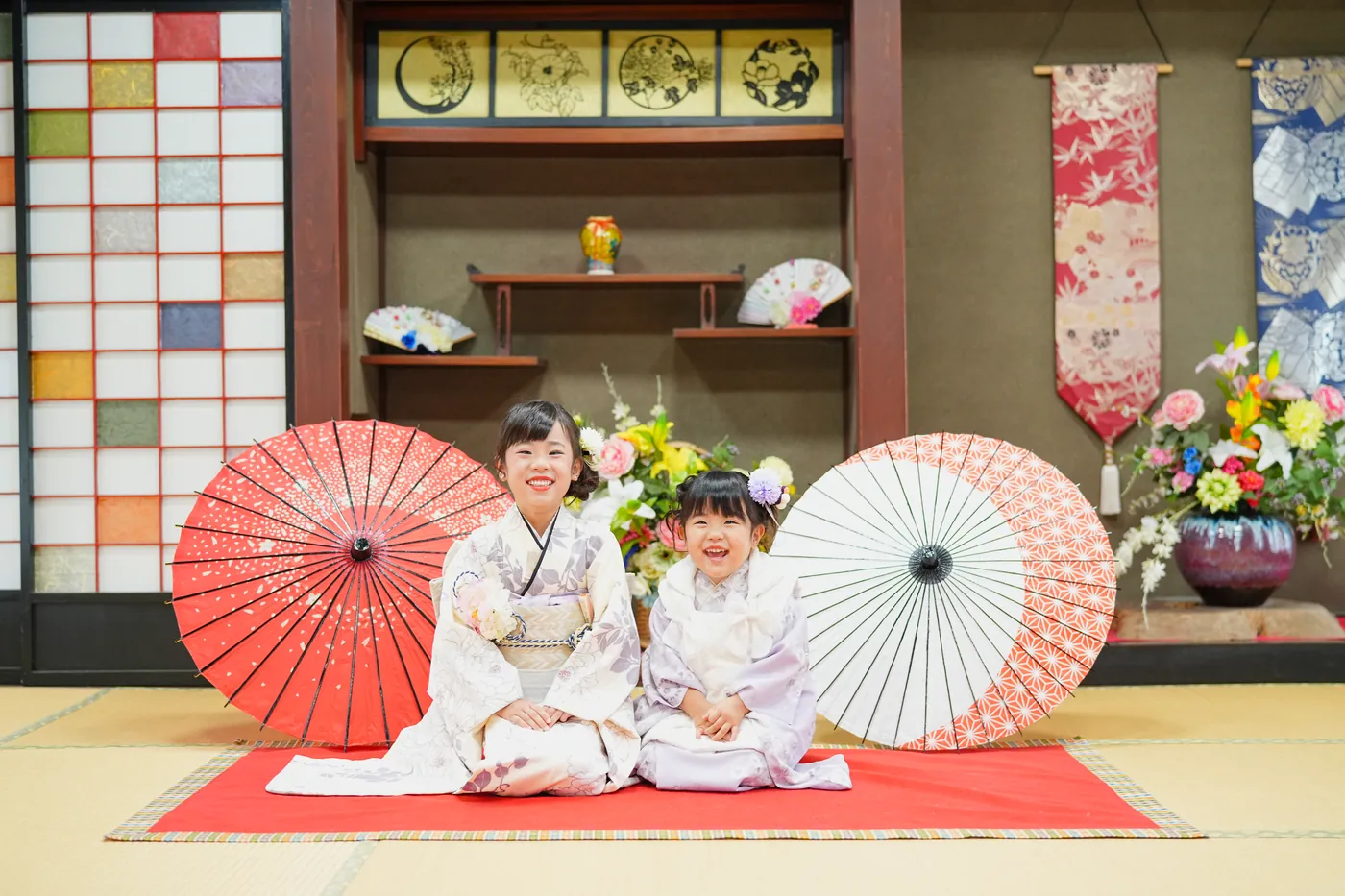 古民家の和室で赤と白の和傘の間に座る着物の姉妹